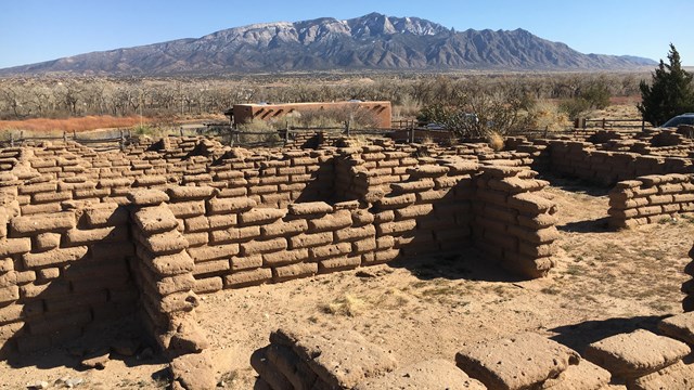 Ruins of adobe brick walls lead to a distant view of a high desert mountain capped with snow.